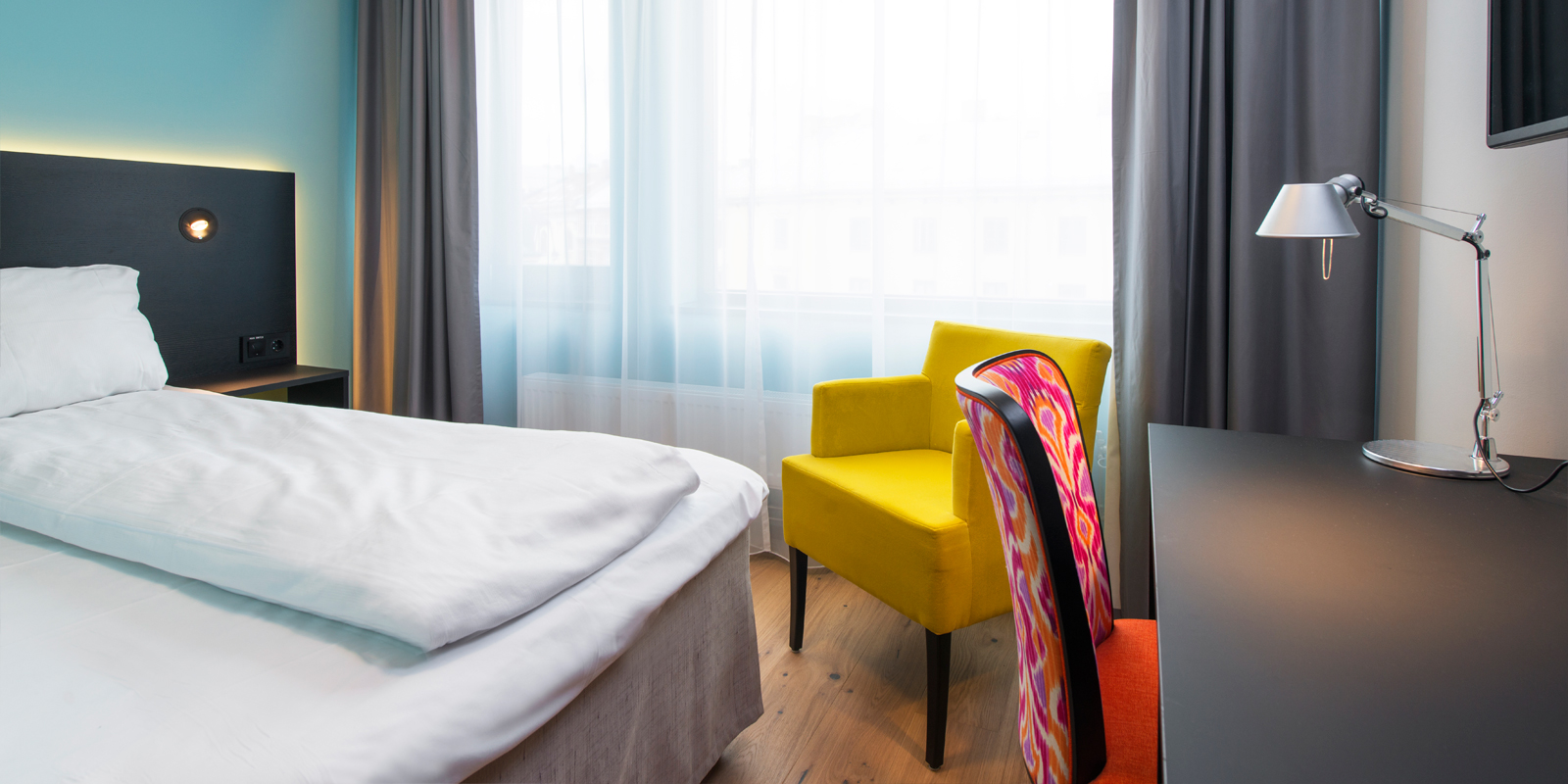 standard room single enkeltseng, nattbord med leselys, en lenestol, vindu med lystette gardiner, skrivebord med lampe, parkettgulv