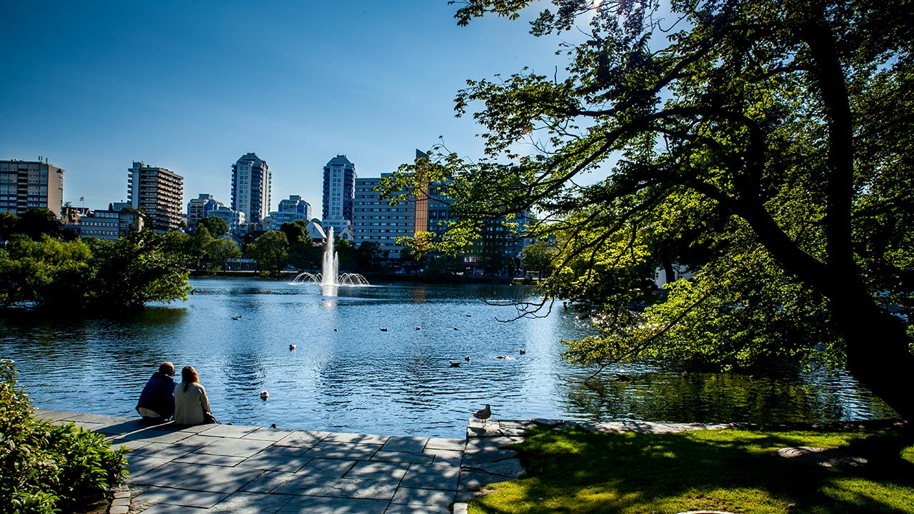 To personer sitter på en benk ved en innsjø med en bysilhuett i bakgrunnen.