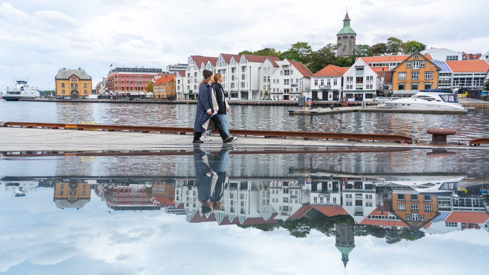 To kvinner som går på en brygge med bygninger i bakgrunnen.
