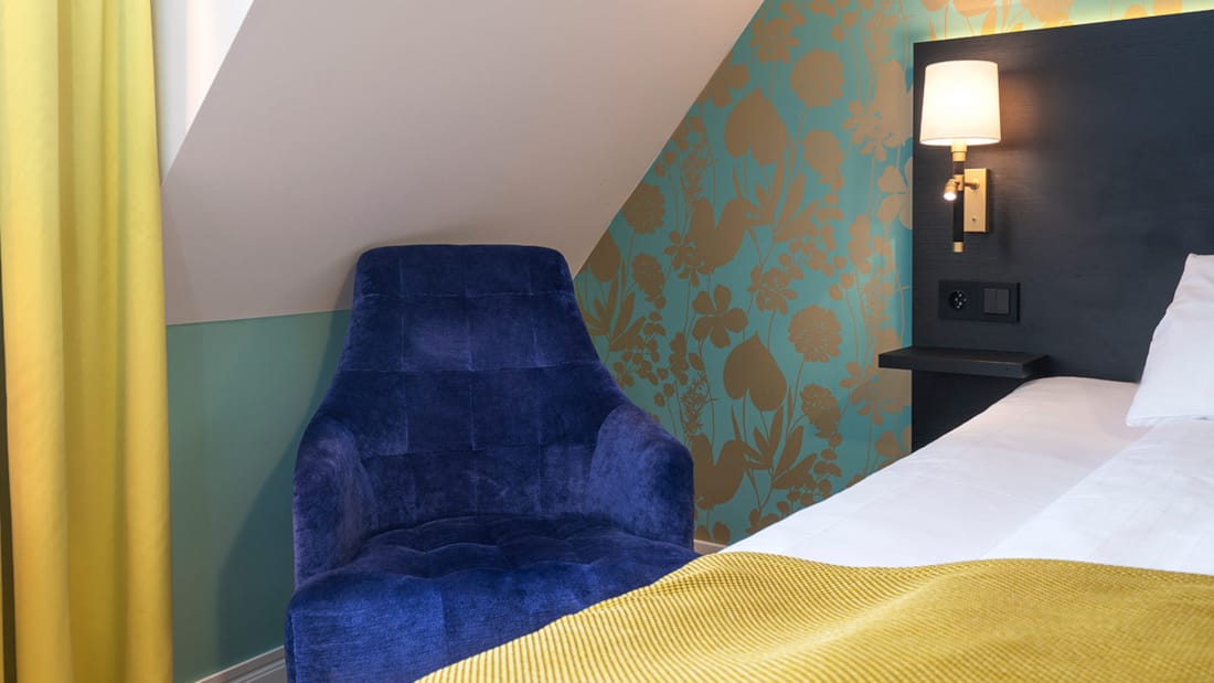 Hjørnet av et standard rom på Thon Hotel Nidaros med en blå stol og kanten på en oppredd seng med gyldent laken og en påslått lampe