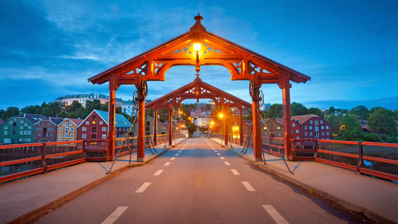 Bilde av Gamle Bybro i Trondheim. Asfaltert gangbro med to røde portaler i hver ende av broa. Rødt rekkverk på hver side. I bakgrunnen ses Bakklandet.