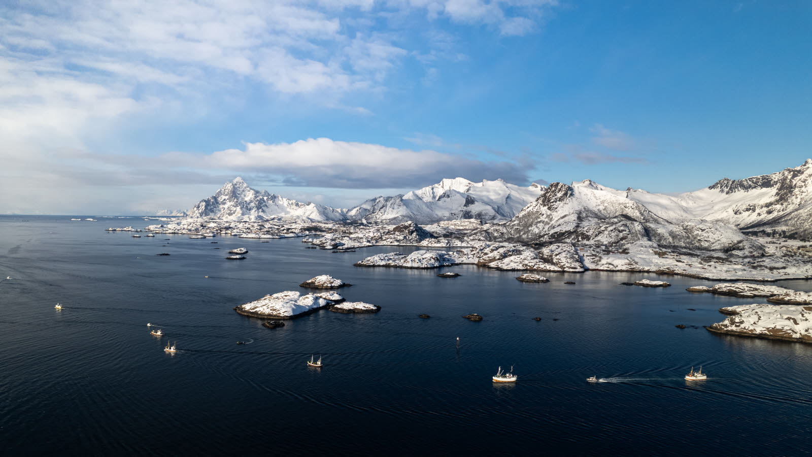 Mange fiskebåter på vannet. I bakgrunnen sees snødekte fjell.