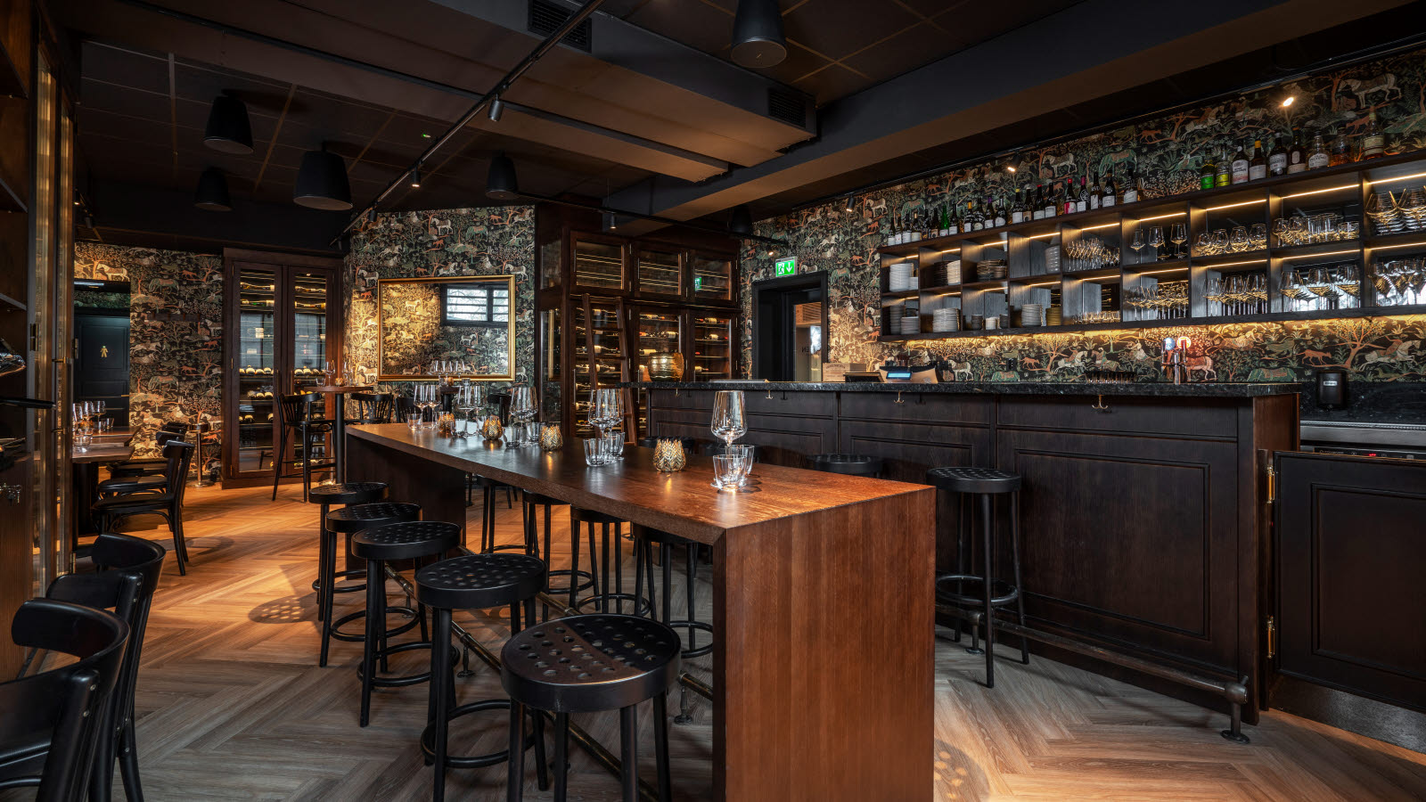 Grotten øl- og vinbar med bardisk med tilhørende runde barkrakker, mørk tapet med tegning av trær, hester og andre dyr og planter, langbord med tilhørende mørke krakker, , høye barbord, parkettgulv. I bakgrunnen ses speil  og vinskap.