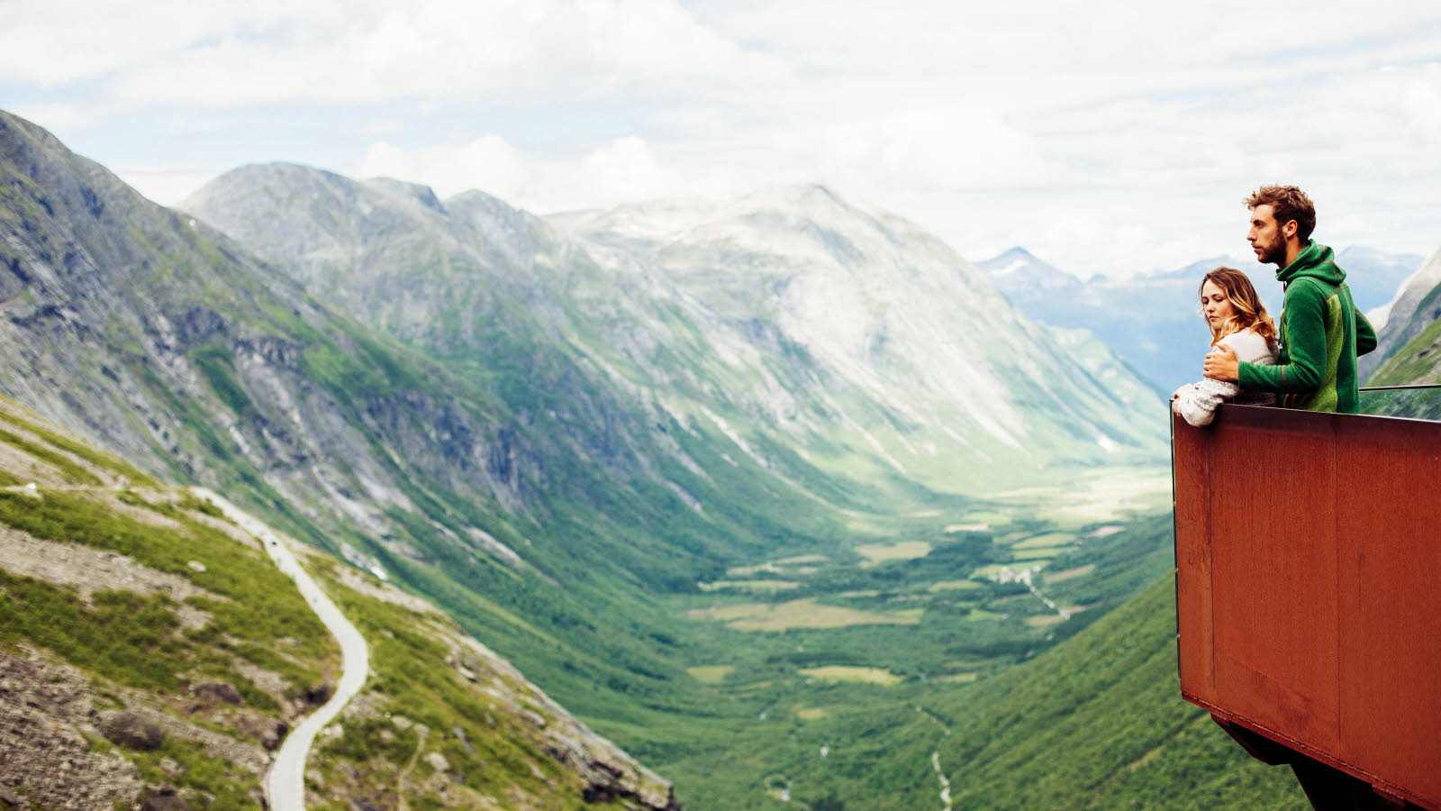 Et par står på utsiktspunktet på toppen av Trollstigen og ser utover veien som snirkler seg oppover.