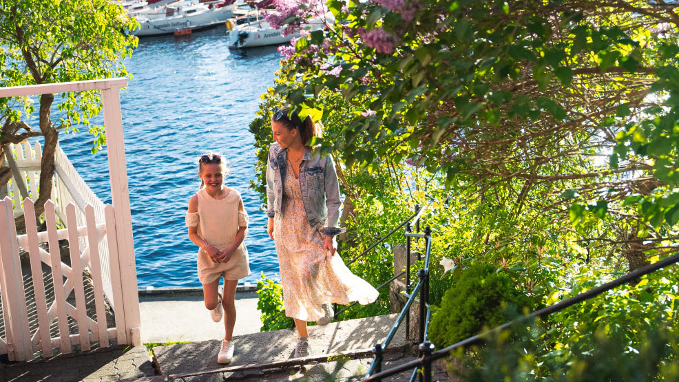 Mor og datter går opp en trapp ute i sommerlige omgivelser.