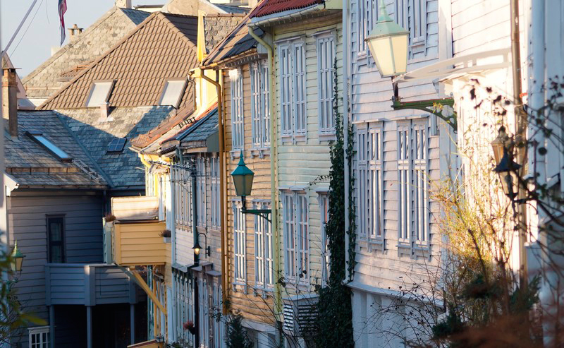 Gammel trehusbebyggelse i Bergen sentrum med planter langs kantene og solen som skinner på veggene.