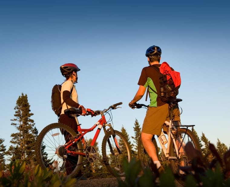 To syklister med hjelm og sekk sett fra et froskeperspektiv. Den ene står ved siden av en rød sykkel, den andre står med ett ben på pedal og det andre benet på andre siden av sykkelen. 
