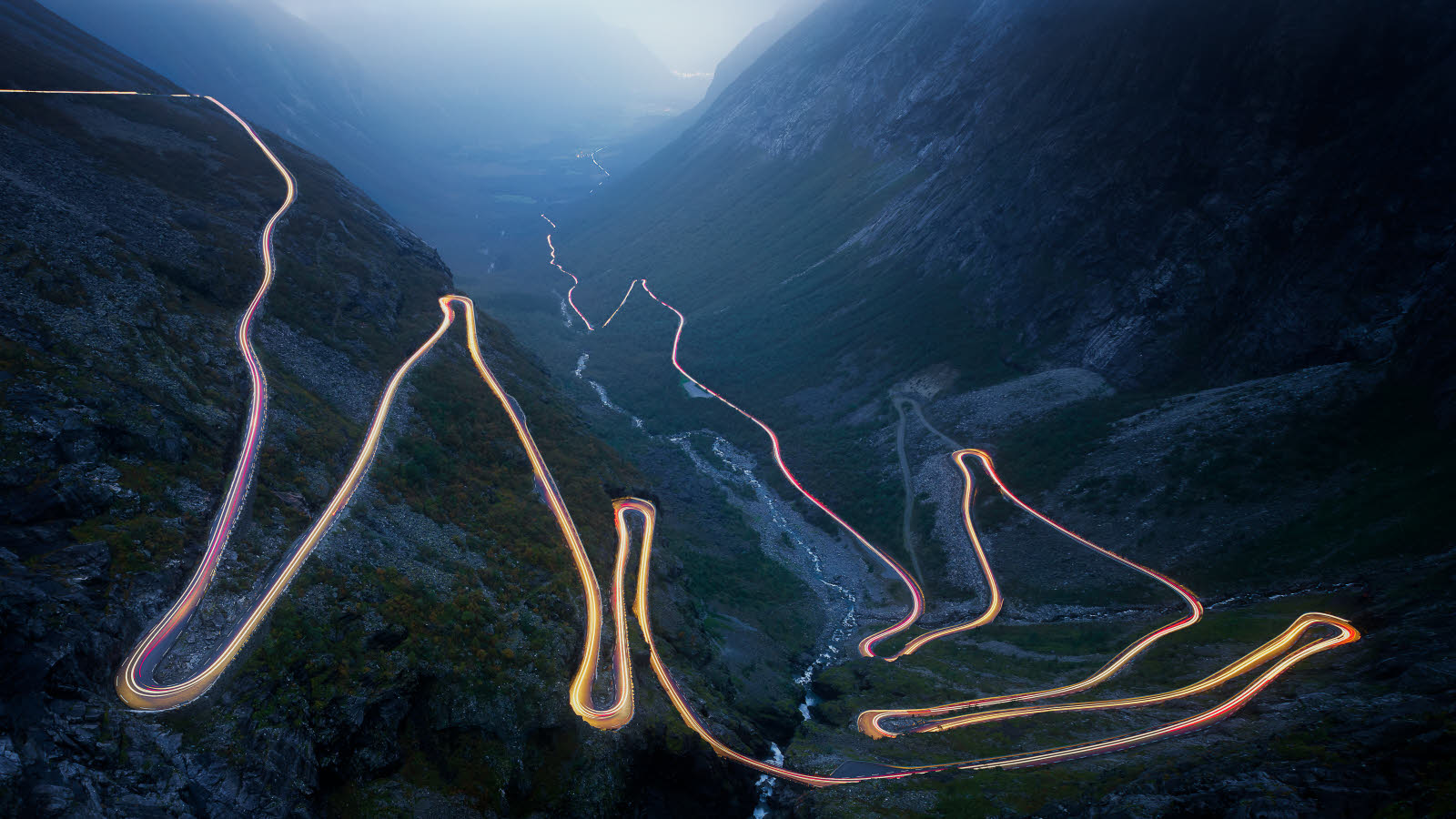 Bilde tatt på kveldstid med lang lukkertid. Trollstigen er lyst opp av gule og røde billys hele veien. I bakgrunnen ses fjell i skumring.