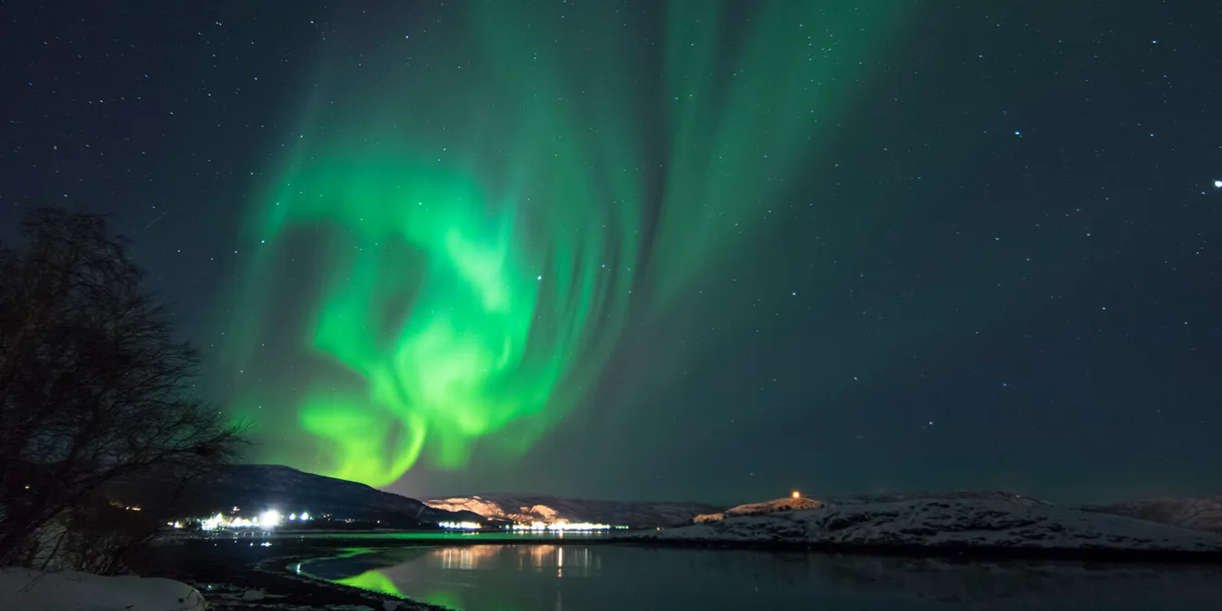 Dansende gr&oslash;nt nordlys over himmelen med en opplyst by og fjellandskap i horisonten.