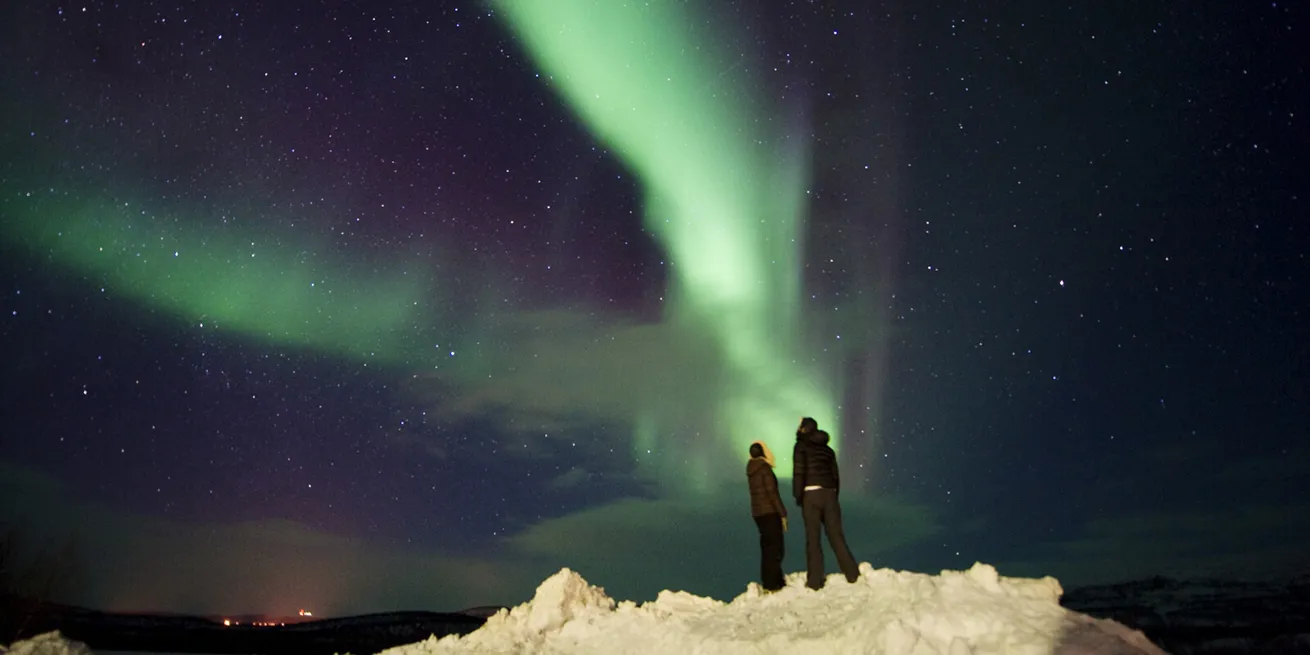 To mennesker st&aring;r p&aring; en sn&oslash;haug og ser opp mot dansende gr&oslash;nt nordlys p&aring; en stjerneklar himmel.