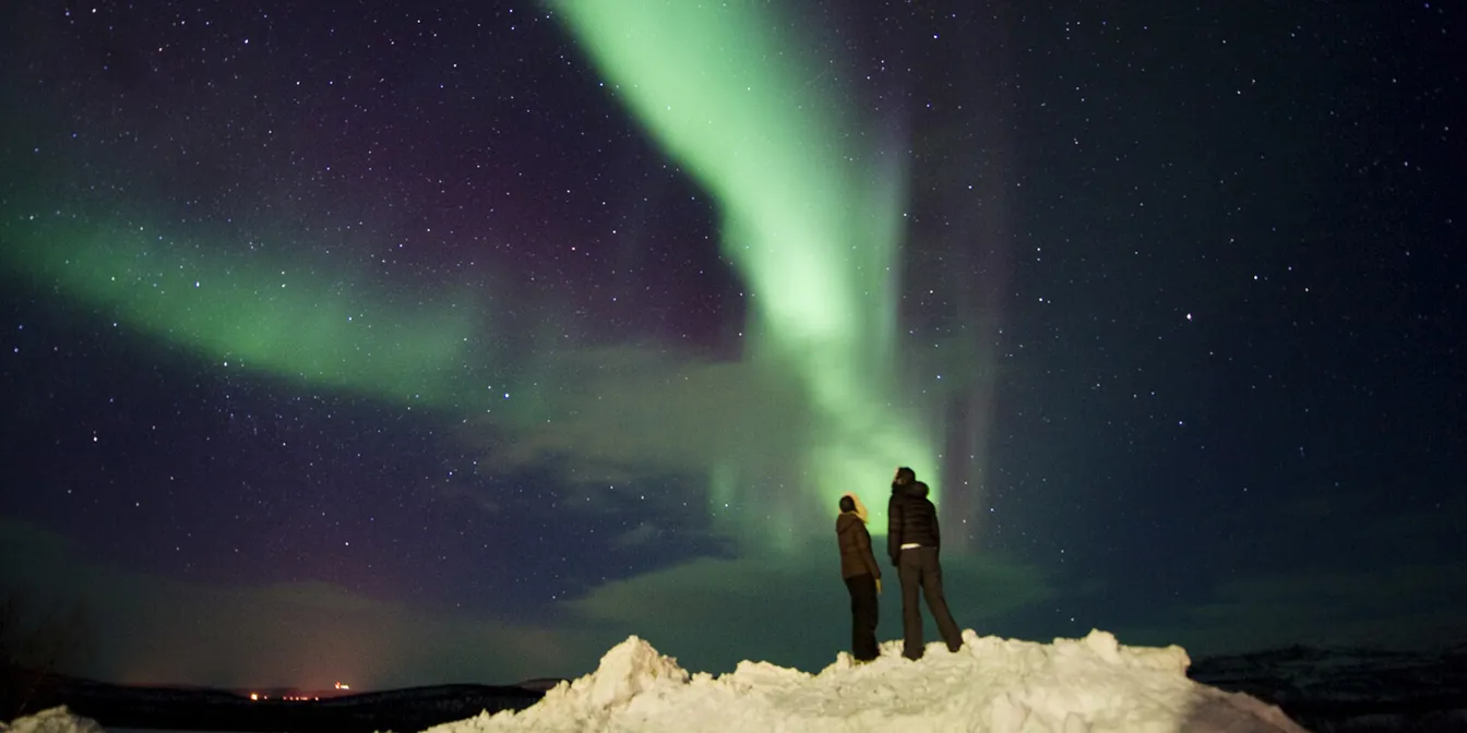 To mennesker st&aring;r p&aring; en sn&oslash;haug og ser opp mot dansende gr&oslash;nt nordlys p&aring; en stjerneklar himmel.