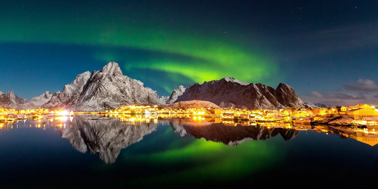 Dansende nordlys i Lofoten over fjell og lysende by i m&oslash;rket som gir gjenskinn i havet i front.