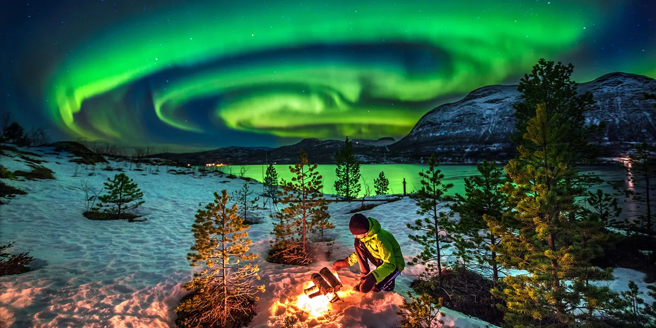 En person som sitter p&aring; huk og tenner opp b&aring;l i et vinterlandskap langs et vann med snirklende gr&oslash;nt nordlys p&aring; himmelen.