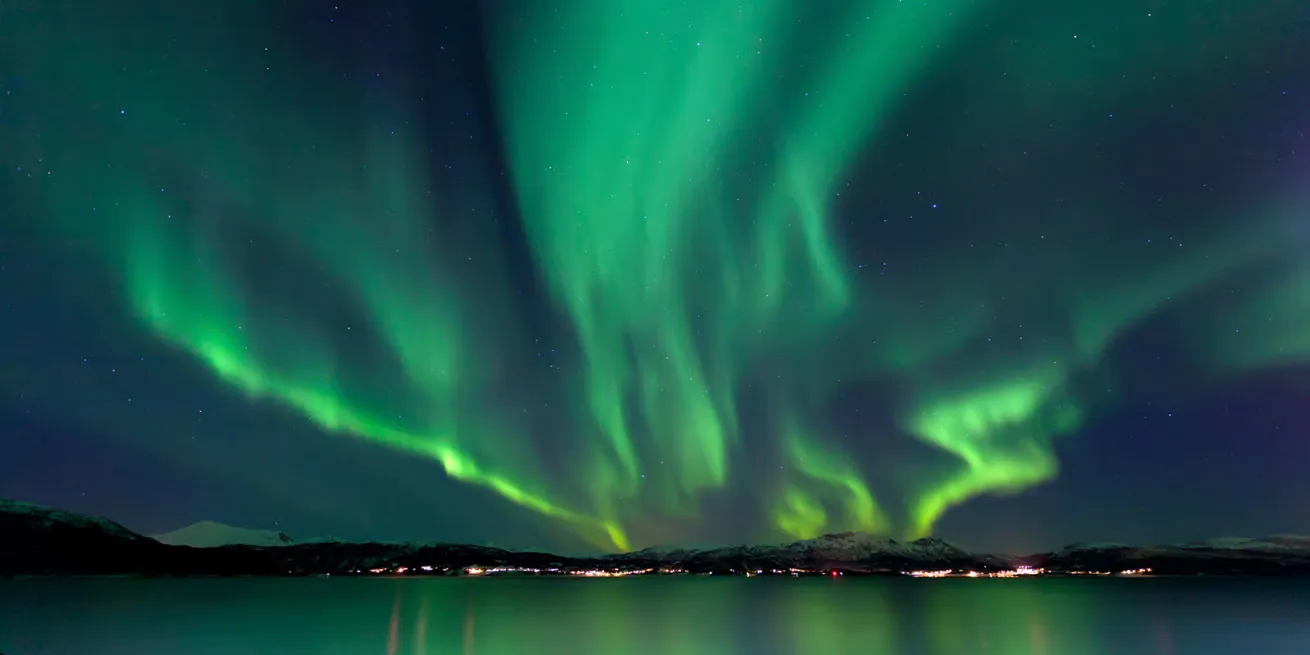 Dansende gr&oslash;nt nordlys p&aring; himmelen over en fjellkjede og bylys i horisonten.