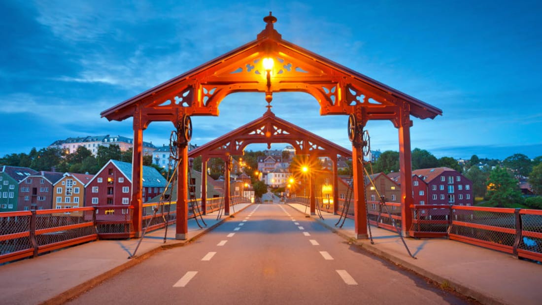 Gamle Bybro i Trondheim. Asfaltert gangbro med to røde portaler i hver ende av broa. Rødt rekkverk på hver side. I bakgrunnen ses Bakklandet.