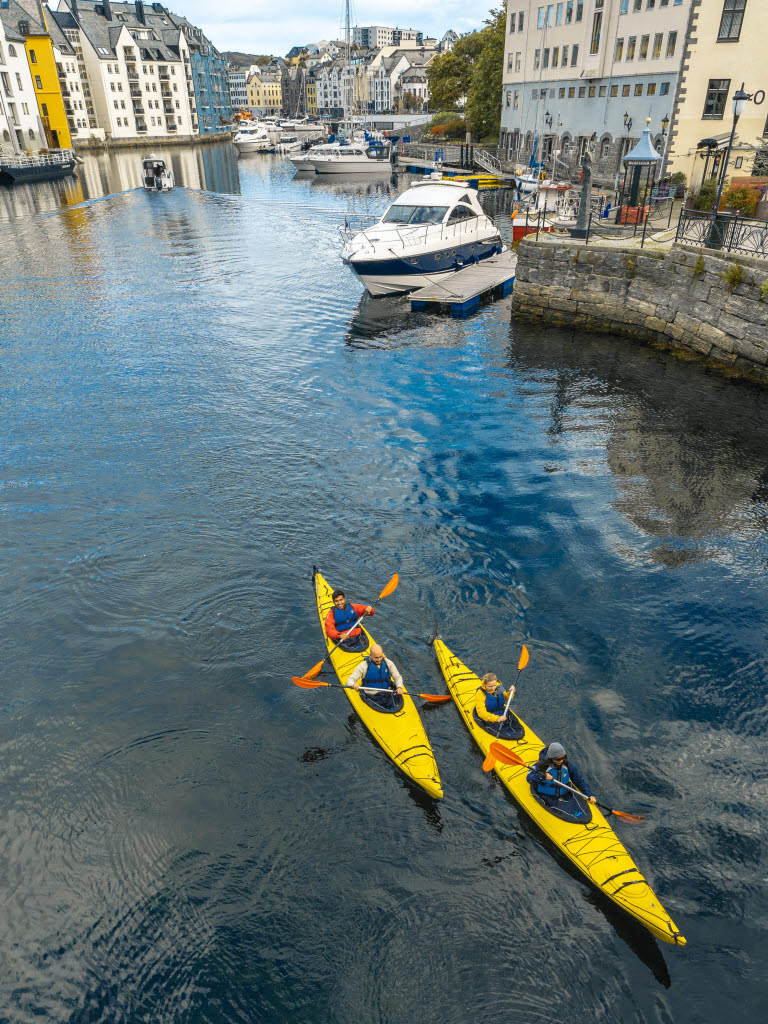 To kajakker med 4 personer på vei ut fra Ålesund sentrum, i Brosundet. I bakgrunnen ses Ålesund sentrum.