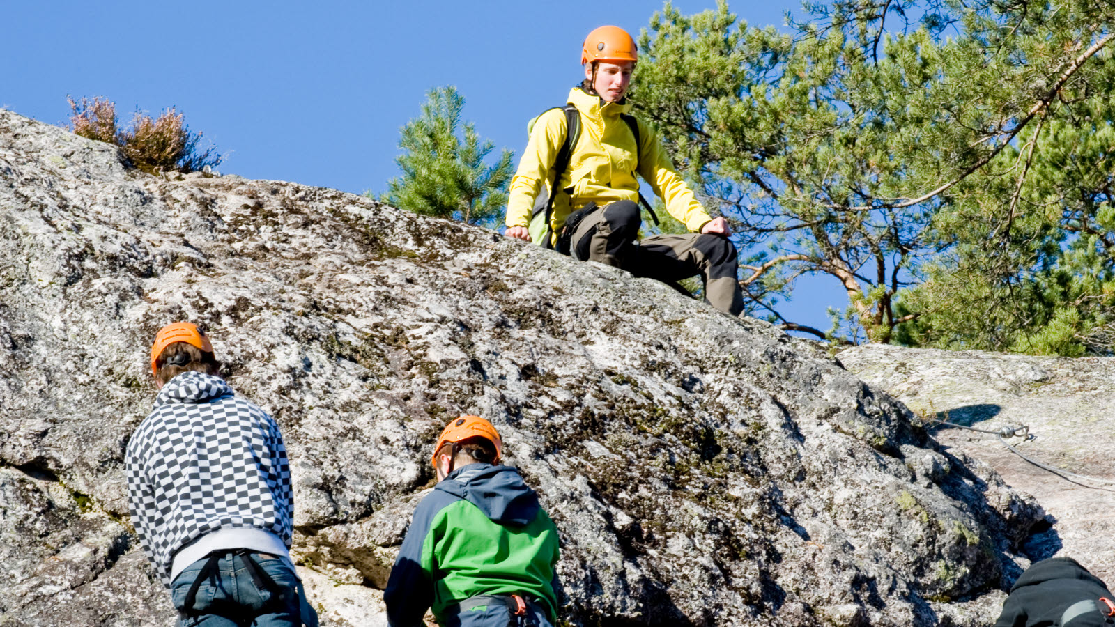 Tre personer iført gule jakker klatrer opp en stein.