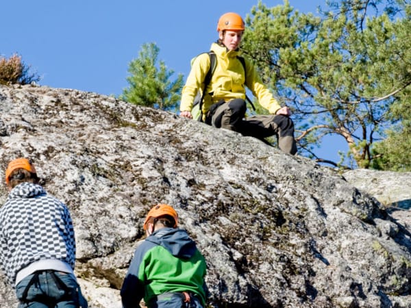 Tre personer iført gule jakker klatrer opp en stein.