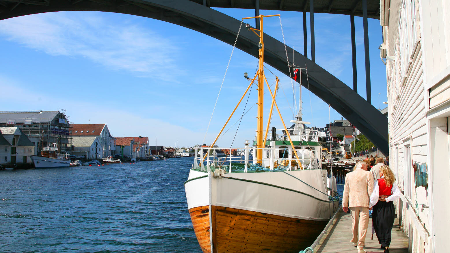 Kvinne og mann som går under en bro med en båt fortøyd i vannet.