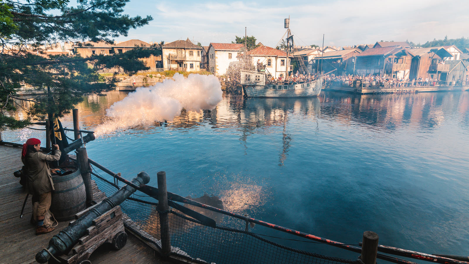 En pirat skyter med kanon over vannet i Dyreparken. På andre siden av vannet ses ei skute, bygninger og masse publikum på brygga.