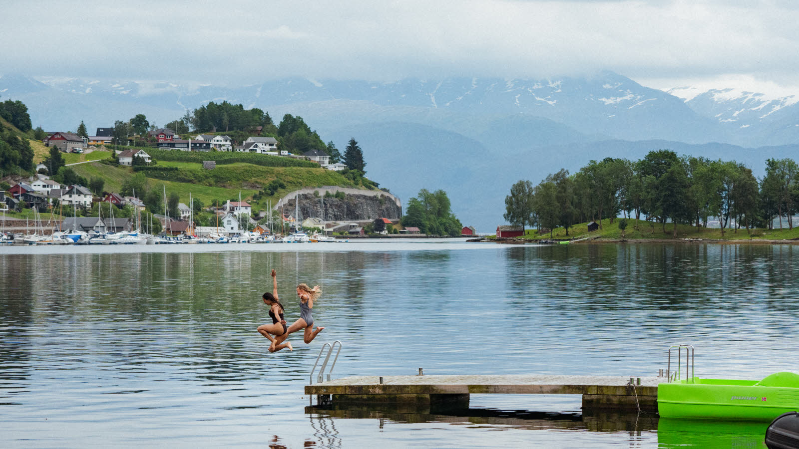 To kvinner hopper i vannet fra en flytebrygge i Norheimsund. I bakgrunnen ses småbåthavn, bebyggelse og fjell med snø på toppen.