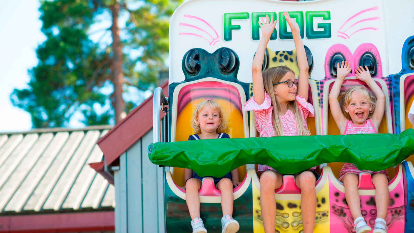 Tre barn sitter i karusellen Frog. To av barna har henda i været.