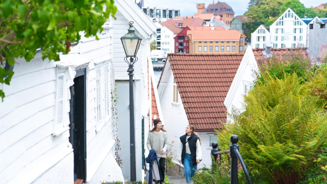 To jenter spaserer i brosteinsbelagt gågate bland hvite huse. I bakgrunnen ses deler av Stavanger sentrum.