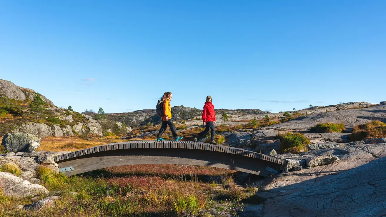 To kvinner som går over en bru ved Preikestolen, sett fra siden. Blå himmel og høstlandskap i bakgrunnen.