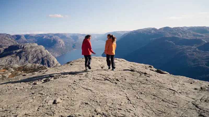 To kvinner går mot kanten av Preikestolen. I Bakgrunnen ses Lysefjorden fra oven, omringet av høye fjell.