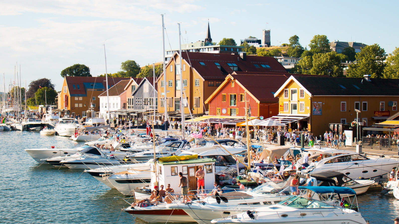 Brygga i Tønsberg en solskinnsdag på sommeren. Langs brygga ligger det båter og seilbåter i ulike størrelser, med mennesker, i flere rader. Til venstre ses Thon Hotel Tønsberg Brygge. Mot høyre ses rekke med restauranter. I bakgrunnen ses Slottsfjell.