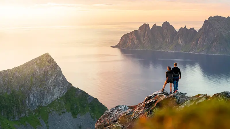 To mennesker som står på et fjell i solnedgang og ser utover vann- og fjellandskap i Tromsø