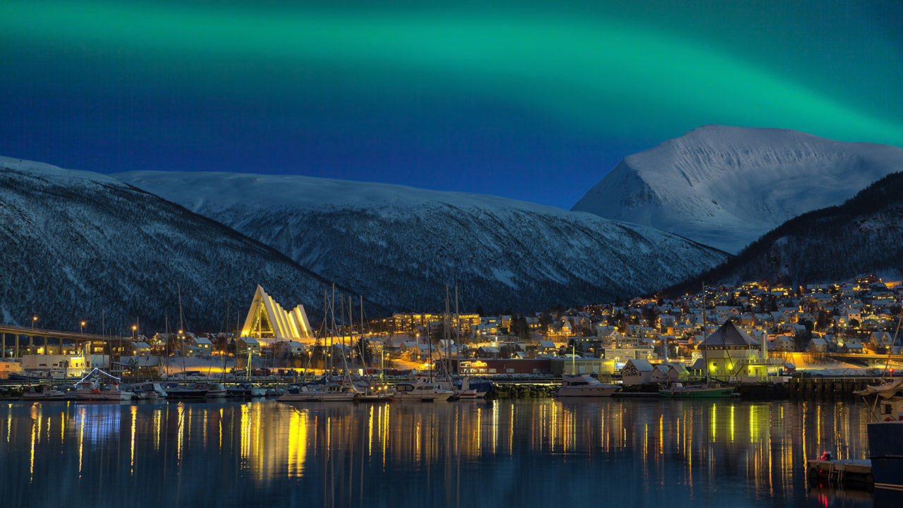 Dansende grønt nordlys på himmelen over Tromsø by som lyser opp i mørket og gir gjenskinn i havet i front.