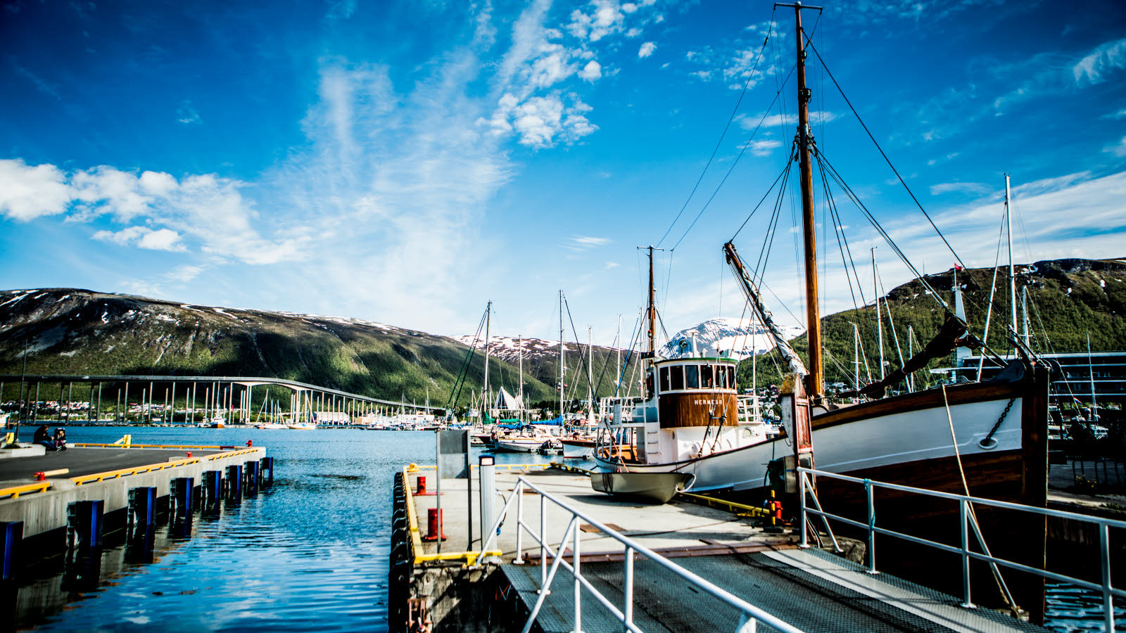 En fiskebåt ligger fortøyd ved en av bryggene i Tromsø en sommerdag. Grønnkledde fjel, Tromsøbrua og Ishavskatedralen ses i bakgrunnen.