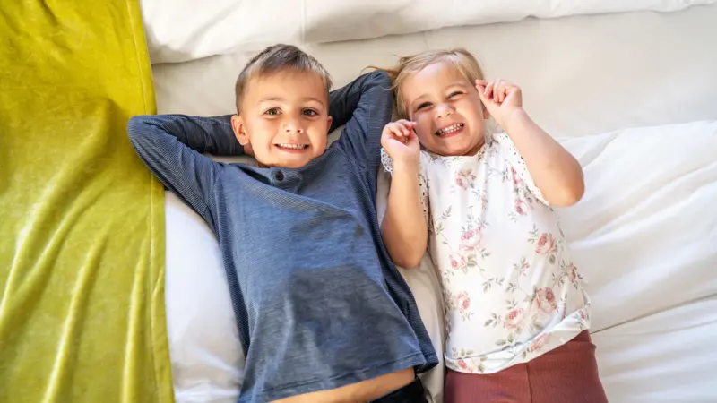 En jente og en gutt som ligger ved siden av hverandre i en hotellseng og smiler til kamera, sett ovenfra.