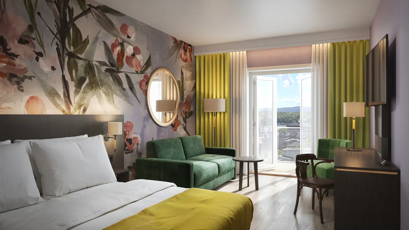 Hotellrom med stor seng og en grønn sofa inntil en vegg med blomstrete tapet i fine farger. På andre siden ser du et skrivebord med en grønn lenestol og et stort vindu med nydelig utsikt ut mot en storby.