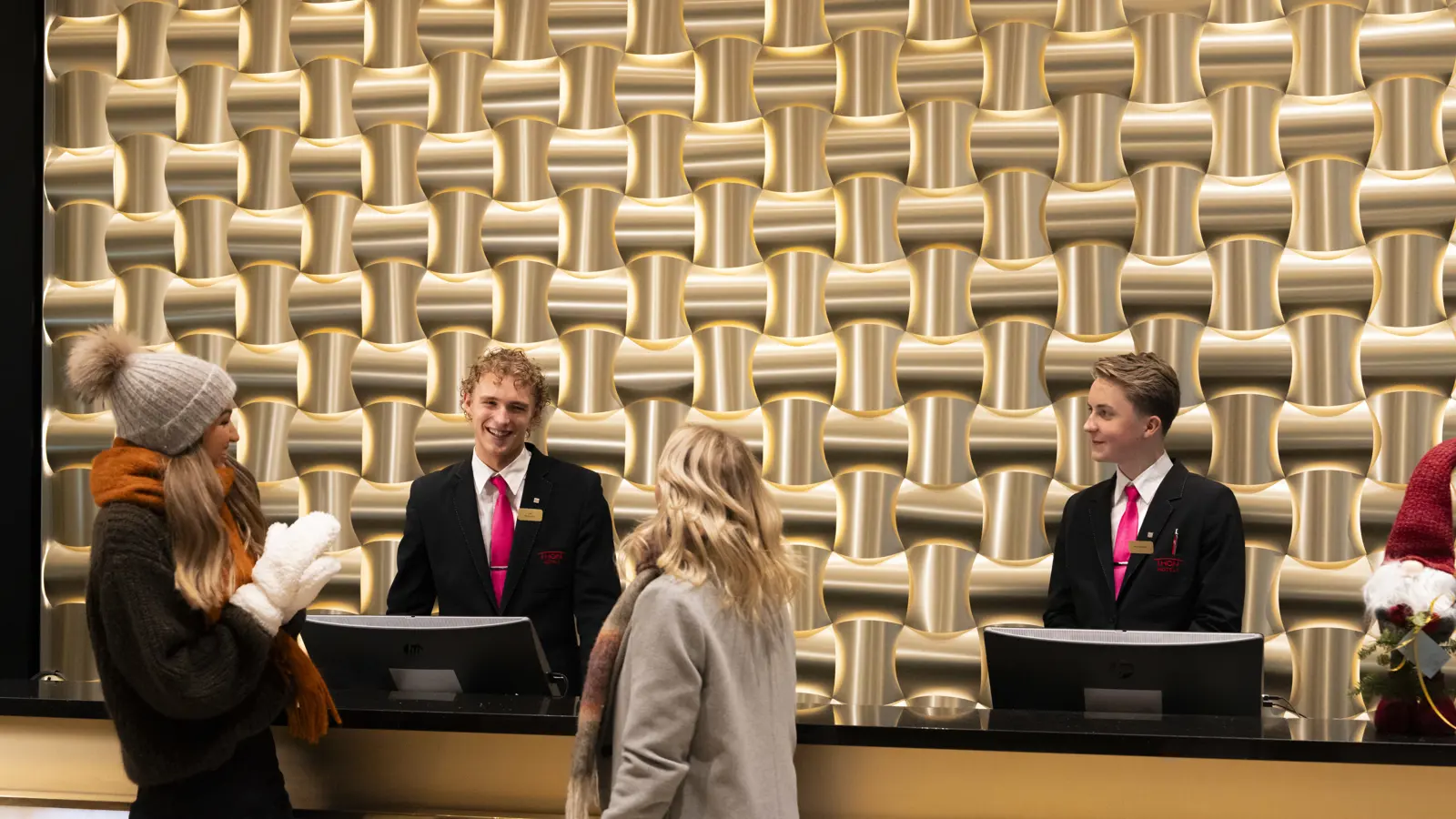 To kunder står foran en hotellresepsjon som er pyntet til jul. De har på seg kåper og luer og smiler mot to ansatte i resepsjonen som hjelper de.