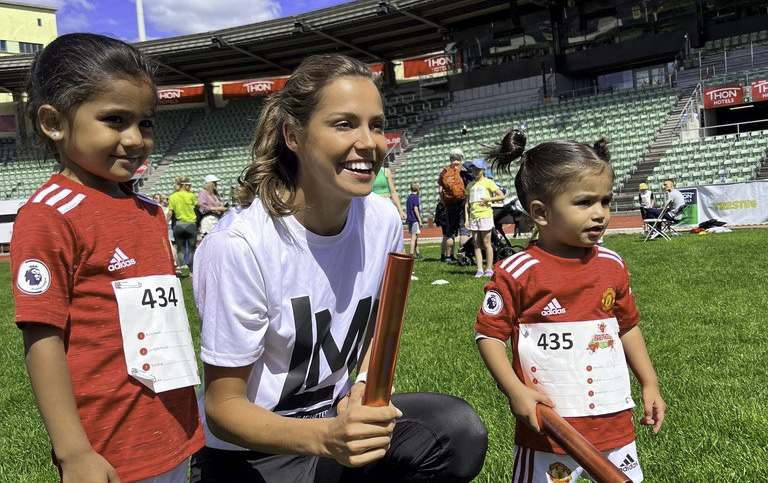 Smilende kvinne på huk med to barn i fotballdrakt stående på hver sin side. Friidrettsarena i bakgrunnen.