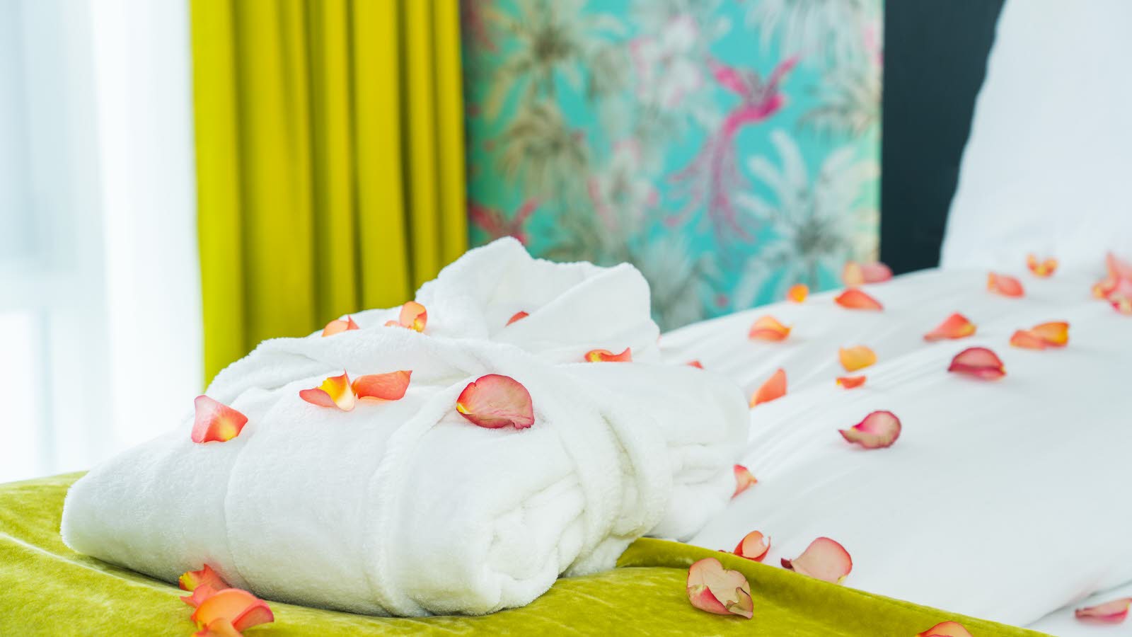 En dobbeltseng med oppredde dyner med hvitt sengetøy hvor det ligger roseblader over sengen og dynene. Gule gardiner, gult sengeteppe og en mønstrete blå turkis tapet i bakgrunnen.
