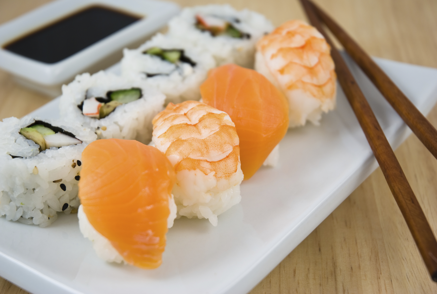 Sushi med laks og ris som ligger dandert på et hvit fat på et bord i eik med spisepinner i mørkt tre ved siden av sushien.