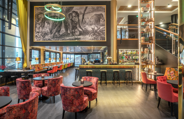 En restaurant med høy takhøyde, kunst på veggen, røde lenestoler og en bar med barkrakker.