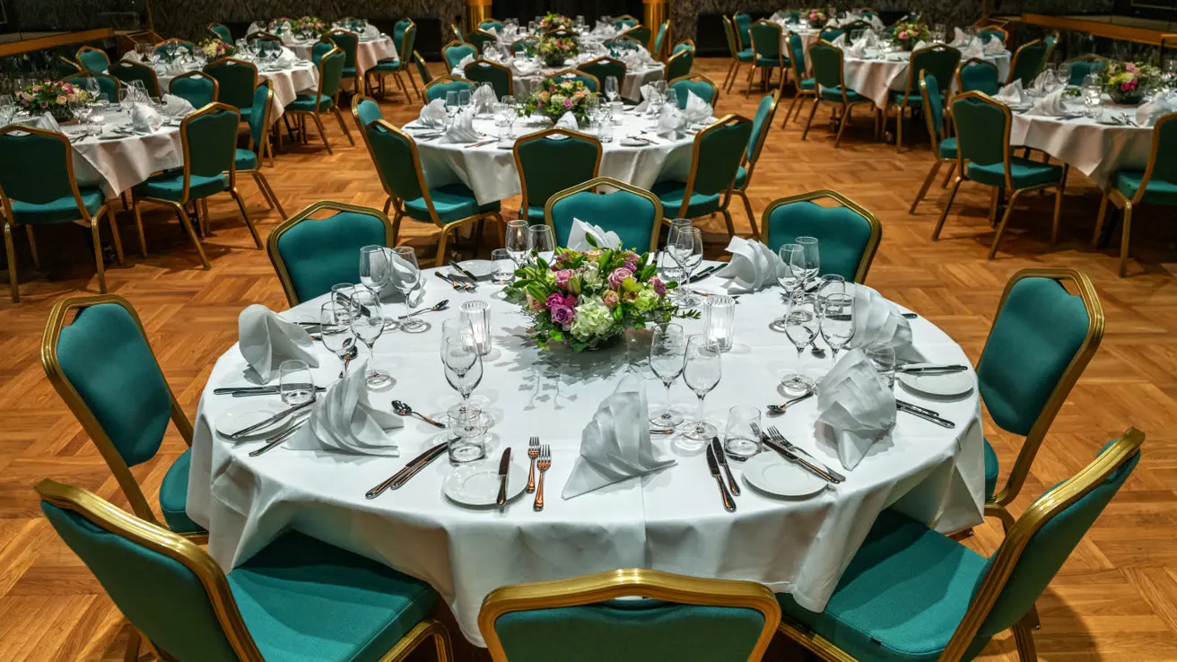Flagstadsalen selskapsoppsett med runde bord og grønne stoler. Bord trukket med hvit duk og oppsett til 8 personer med blomsterbukett midt på bordet.