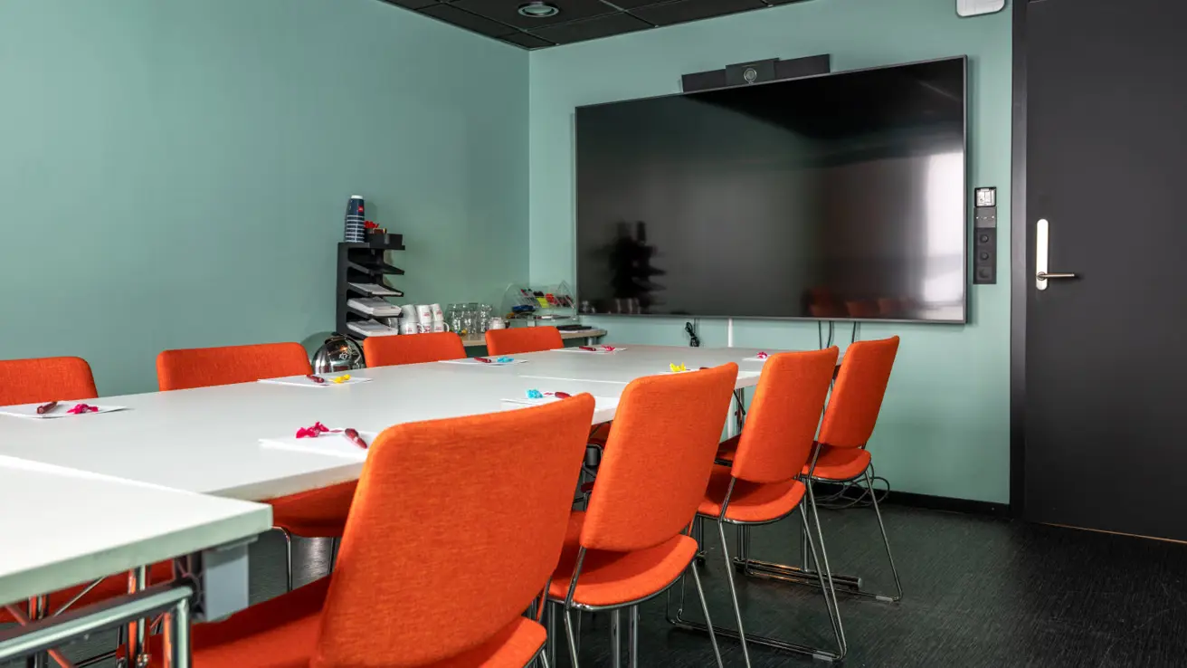 Møterom med langbord og oransje stoler rundt. Lys grønn farge på veggen og stor vegg hengt TV skjerm ved siden av inngangsdør.
