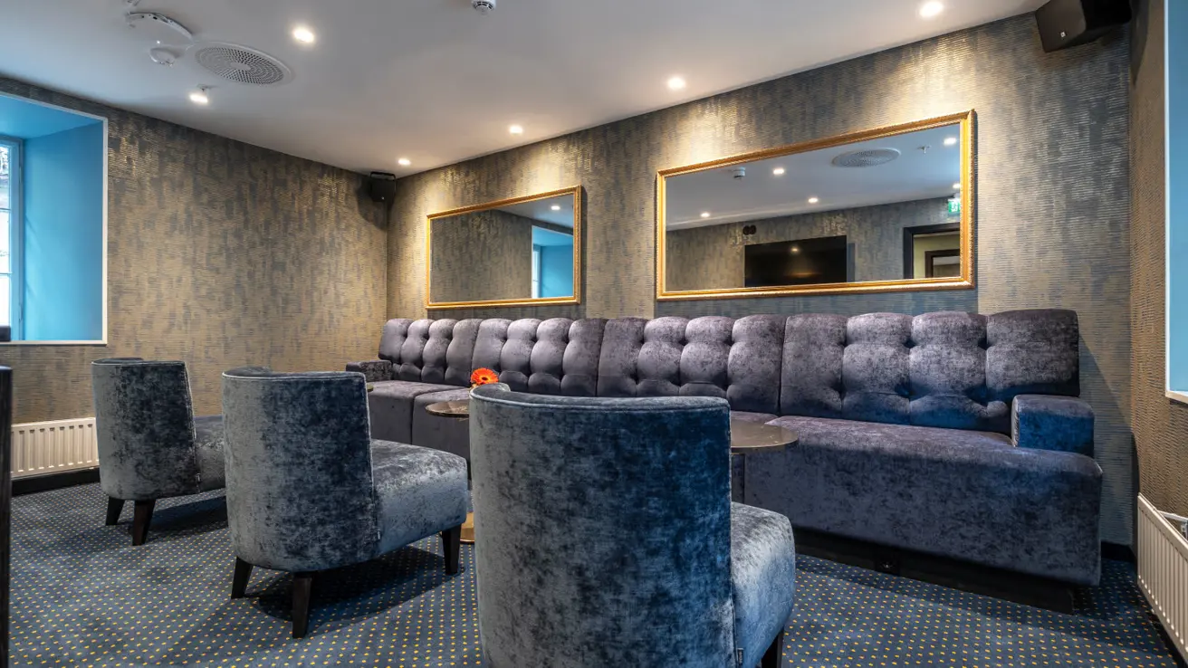 Loungeområde tilknyttet et møterom med blå velour sofaer og lenestoler, speil på veggene og strukturtapet på veggen.