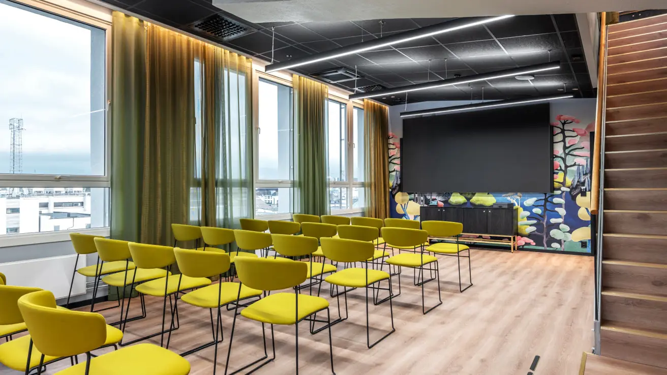 Møteromoppsett med grønne stoler på rekke og rad. Store vinduer langs den ene veggen med utsikt.