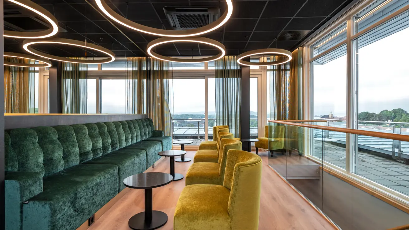 Loungeområde med lang grønn sofa i fløyel og lysegrønne lenestoler, lokalisert i en etasje høyt oppe med store vinduer som gir god utsikt.